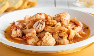 Un invitante piatto di zuppa di pesce surgelato, con calamari, gamberi e pezzi di pesce bianco in un sugo rosso, servito con pane tostato.