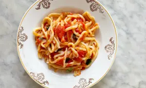 Piatto di pasta con sugo all'aglione e basilico, servito in un piatto bianco con decorazioni dorate su sfondo marmorizzato.