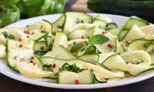 Fette di zucchine marinate, condite con pepe rosa e erbe aromatiche, su un piatto bianco.