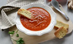 Una ciotola di crema di peperoni, guarnita con crostini e un filo d'olio. Accanto, un cucchiaio, un peperone rosso e zenzero.