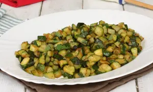Zucchine in padella, tagliate a cubetti e condite con erbe aromatiche fresche, servite su un piatto bianco.