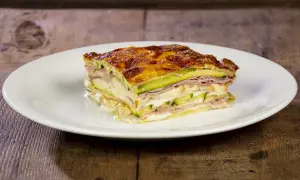 Una fetta di parmigiana di zucchine, stratificata con prosciutto e formaggio filante, dorata al forno su un piatto bianco.