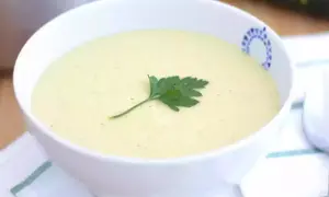 Una cremosa vellutata di zucchine, guarnita con un rametto di prezzemolo fresco, servita in una ciotola bianca con un bordo decorato.
