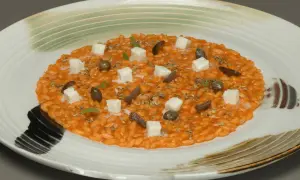 Risotto alla pizzaiola cremoso, guarnito con cubetti di formaggio, olive, capperi e erbe aromatiche su un piatto bianco decorato.
