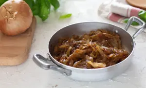 Cipolle caramellate in padella, pronte per insaporire un piatto. Accanto, una cipolla cruda su un tagliere.