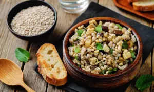 Insalata di farro con verdure miste, pinoli e menta, servita in una ciotola di legno. Accanto, pane tostato e una ciotola di farro crudo.