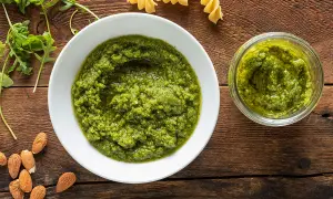 Pesto di rucola e mandorle in una ciotola bianca, accanto a un vasetto pieno, con mandorle e rucola fresca sparse sul tavolo di legno.