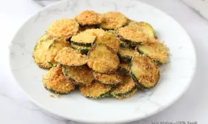 Fettine di zucchine impanate e cotte in friggitrice ad aria, dorate e croccanti, servite su un piatto bianco con venature grigie.