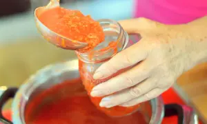 Mani che versano pomarola ricetta in un barattolo di vetro. Il sugo fumante viene preso da una pentola con un mestolo.