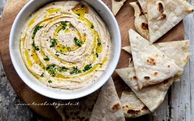 Delizioso hummus di fagioli cremoso, condito con olio d'oliva, prezzemolo e semi di sesamo, servito con pane pita tostato.
