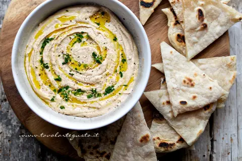 Delizioso hummus di fagioli cremoso, condito con olio d'oliva, prezzemolo e semi di sesamo, servito con pane pita tostato.
