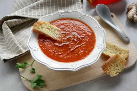 Una ciotola di crema di peperoni, guarnita con crostini e un filo d'olio. Accanto, un cucchiaio, un peperone rosso e zenzero.