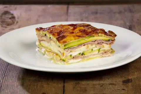 Una fetta di parmigiana di zucchine, stratificata con prosciutto e formaggio filante, dorata al forno su un piatto bianco.
