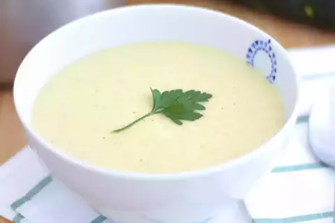 Una cremosa vellutata di zucchine, guarnita con un rametto di prezzemolo fresco, servita in una ciotola bianca con un bordo decorato.