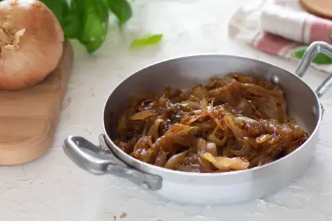 Cipolle caramellate in padella, pronte per insaporire un piatto. Accanto, una cipolla cruda su un tagliere.