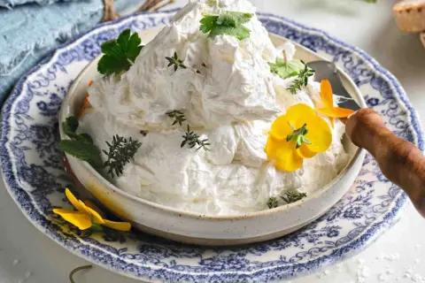Burro montato soffice e arioso, guarnito con erbe aromatiche e fiori gialli, servito in una ciotola decorata.