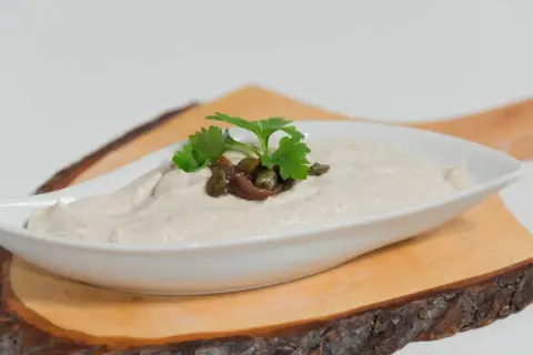 Salsa tonnata cremosa, guarnita con acciughe, capperi e prezzemolo, servita in una ciotola bianca su un tagliere di legno.