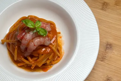 Spaghetti conditi con un sugo rosso intenso, guarniti con gamberi rosa e basilico fresco. Un piatto che evoca il sapore del mare, come una ricca bisque di crostacei.