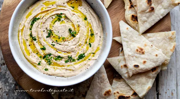 Delizioso hummus di fagioli cremoso, condito con olio d'oliva, prezzemolo e semi di sesamo, servito con pane pita tostato.