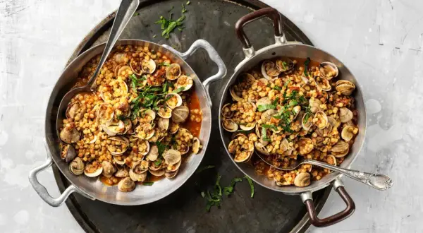 Due padelle piene di fregola con vongole, un piatto sardo profumato, guarnito con prezzemolo fresco.