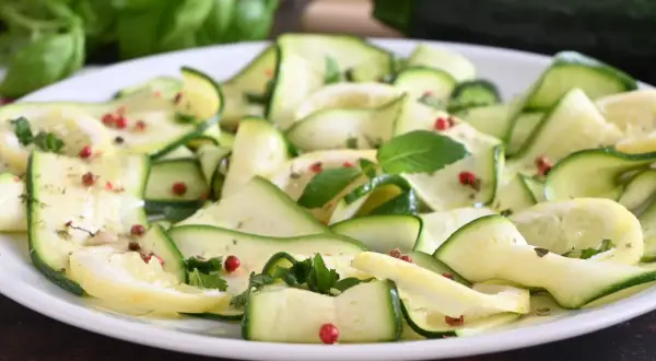 Fette di zucchine marinate, condite con pepe rosa e erbe aromatiche, su un piatto bianco.