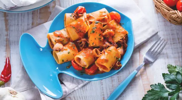 Piatto di pasta con calamaretti, pomodorini e prezzemolo su un piatto a forma di pesce blu.