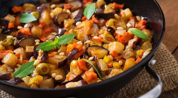 Caponata di verdure in padella, con melanzane, carote, olive e pinoli, guarnita con foglie di basilico fresco.