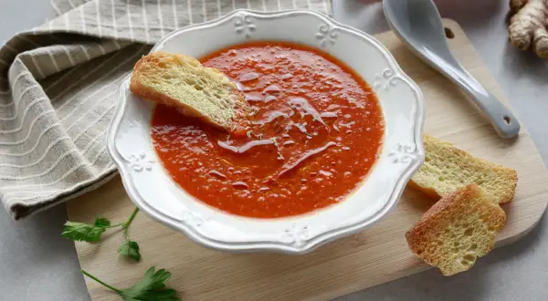 Una ciotola di crema di peperoni, guarnita con crostini e un filo d'olio. Accanto, un cucchiaio, un peperone rosso e zenzero.