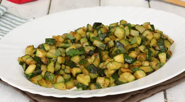 Zucchine in padella, tagliate a cubetti e condite con erbe aromatiche fresche, servite su un piatto bianco.