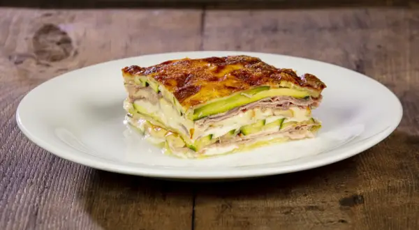 Una fetta di parmigiana di zucchine, stratificata con prosciutto e formaggio filante, dorata al forno su un piatto bianco.