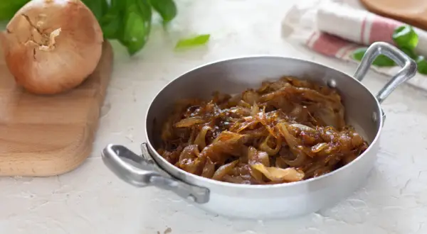 Cipolle caramellate in padella, pronte per insaporire un piatto. Accanto, una cipolla cruda su un tagliere.