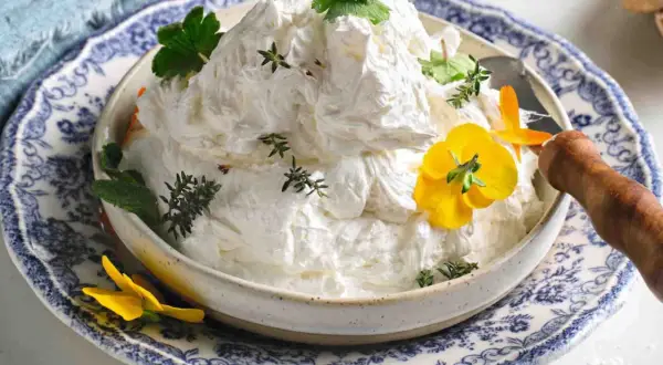 Burro montato soffice e arioso, guarnito con erbe aromatiche e fiori gialli, servito in una ciotola decorata.