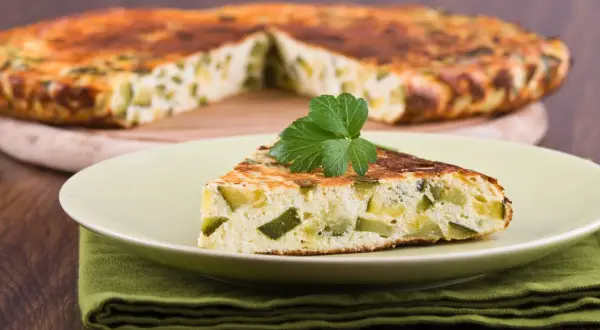 Una fetta di frittata di zucchine, soffice e dorata, guarnita con prezzemolo fresco, su un piatto chiaro.