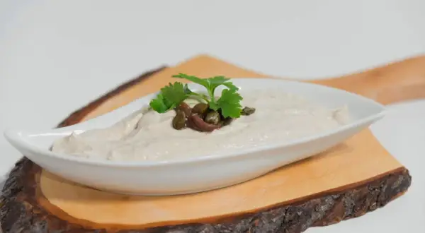 Salsa tonnata cremosa, guarnita con acciughe, capperi e prezzemolo, servita in una ciotola bianca su un tagliere di legno.