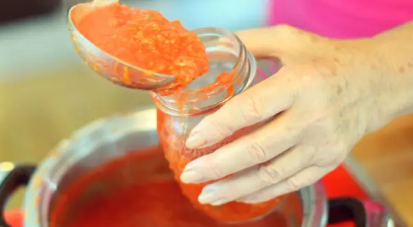 Mani che versano pomarola ricetta in un barattolo di vetro. Il sugo fumante viene preso da una pentola con un mestolo.