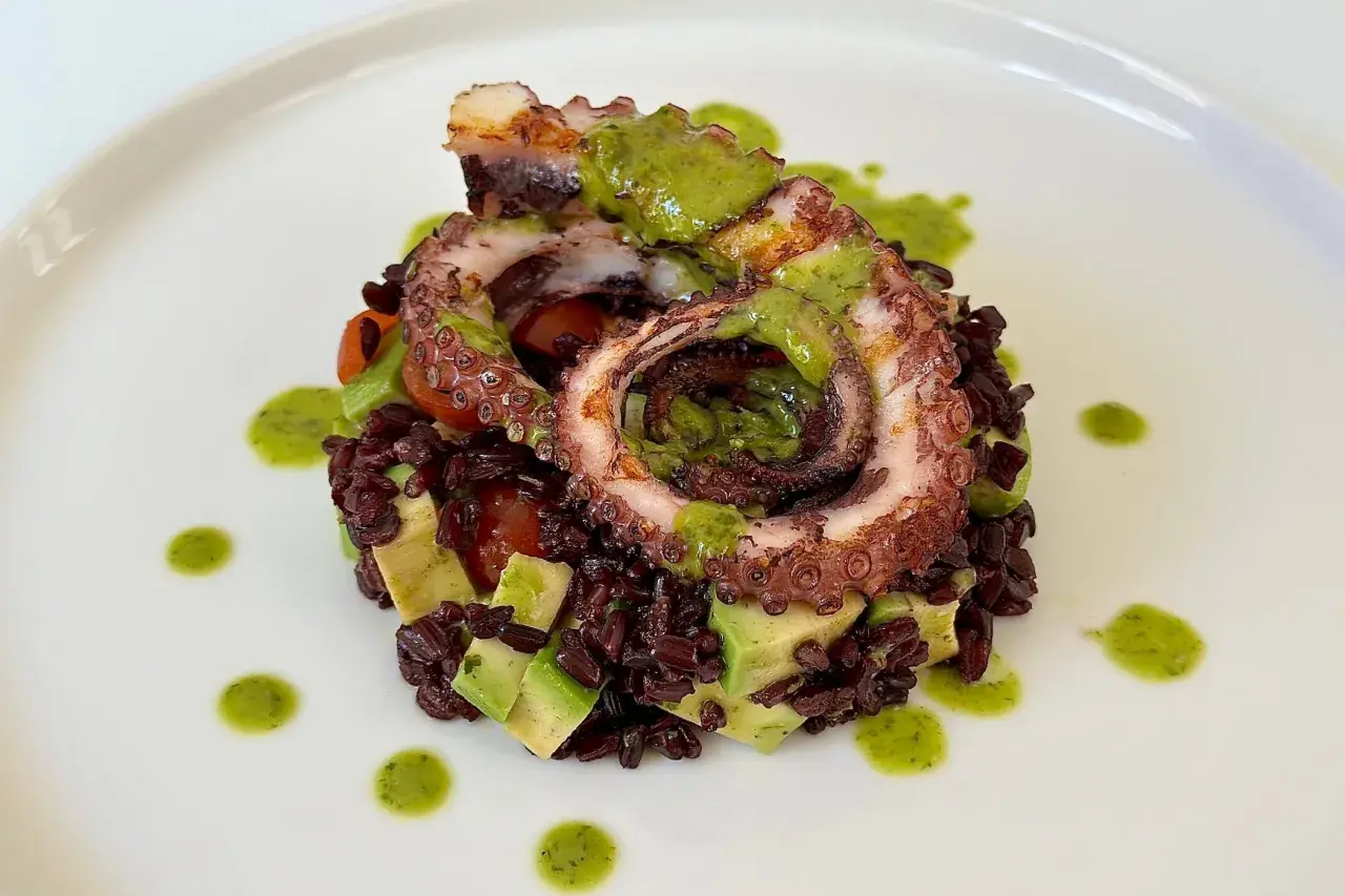 Delizioso polpo alla griglia servito su riso venere con avocado e pomodorini, guarnito con salsa verde.