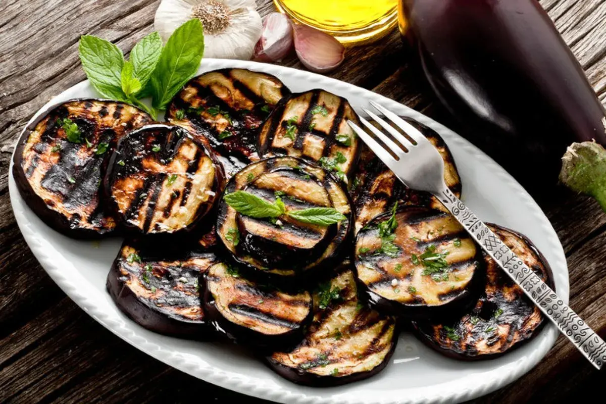 Melanzane grigliate su un piatto bianco, condite con erbe aromatiche. Un invito a scoprire i 100 modi per cucinare le melanzane.