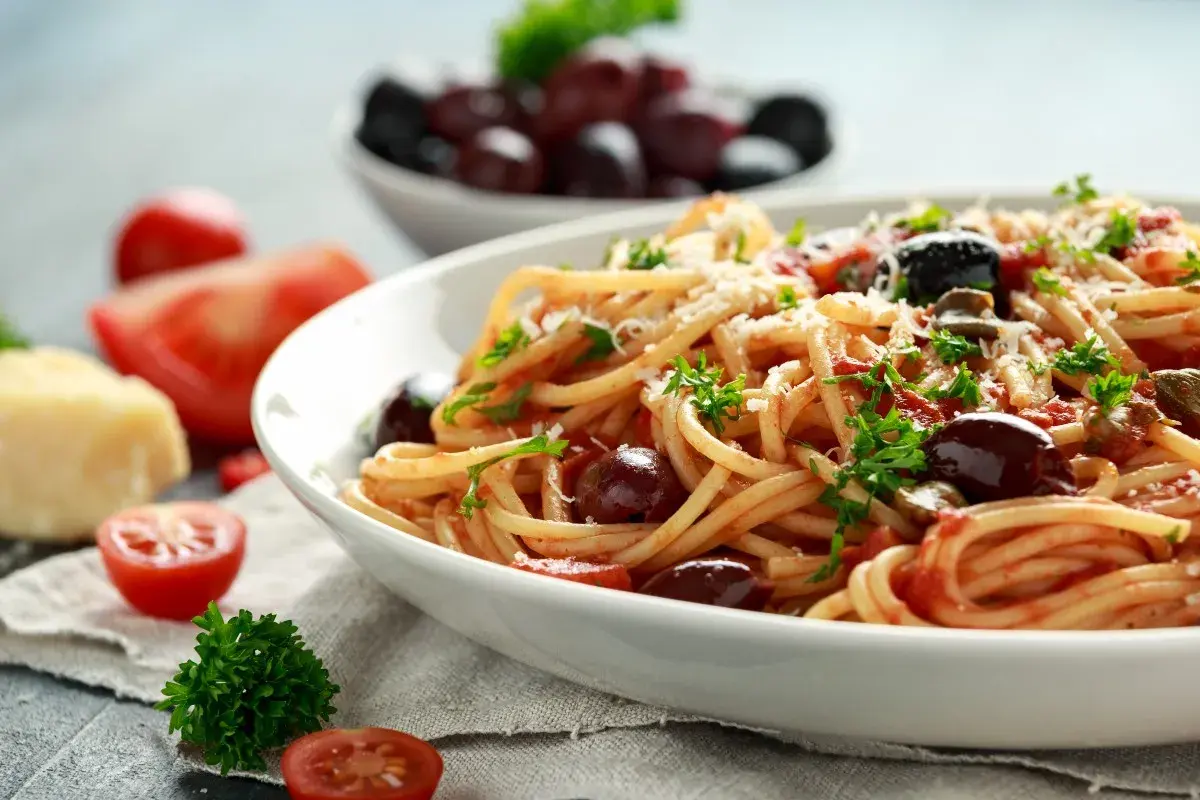 Spaghetti al sugo alla puttanesca, con pomodoro, olive, capperi e prezzemolo fresco, guarniti con parmigiano grattugiato.