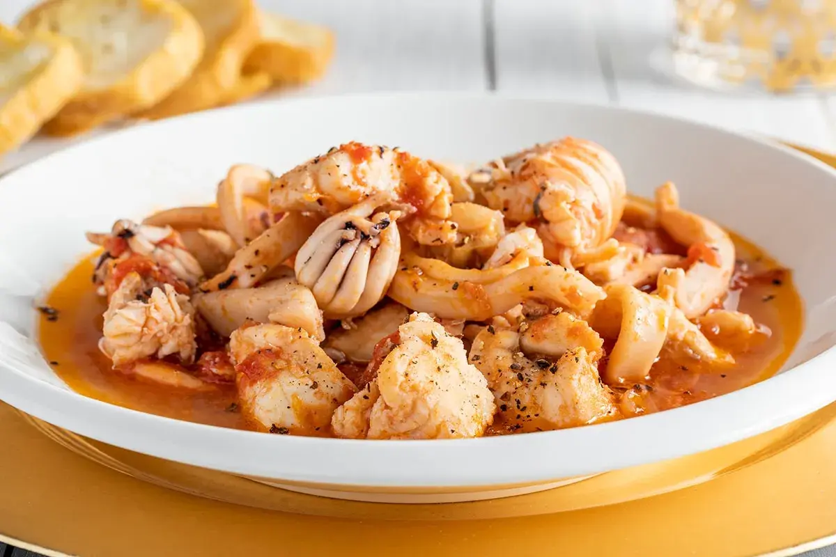 Un invitante piatto di zuppa di pesce surgelato, con calamari, gamberi e pezzi di pesce bianco in un sugo rosso, servito con pane tostato.