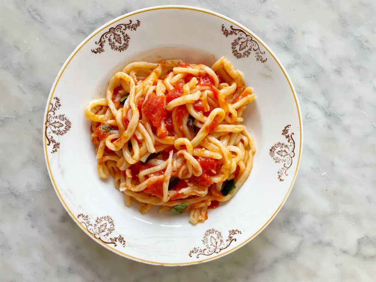 Piatto di pasta con sugo all'aglione e basilico, servito in un piatto bianco con decorazioni dorate su sfondo marmorizzato.