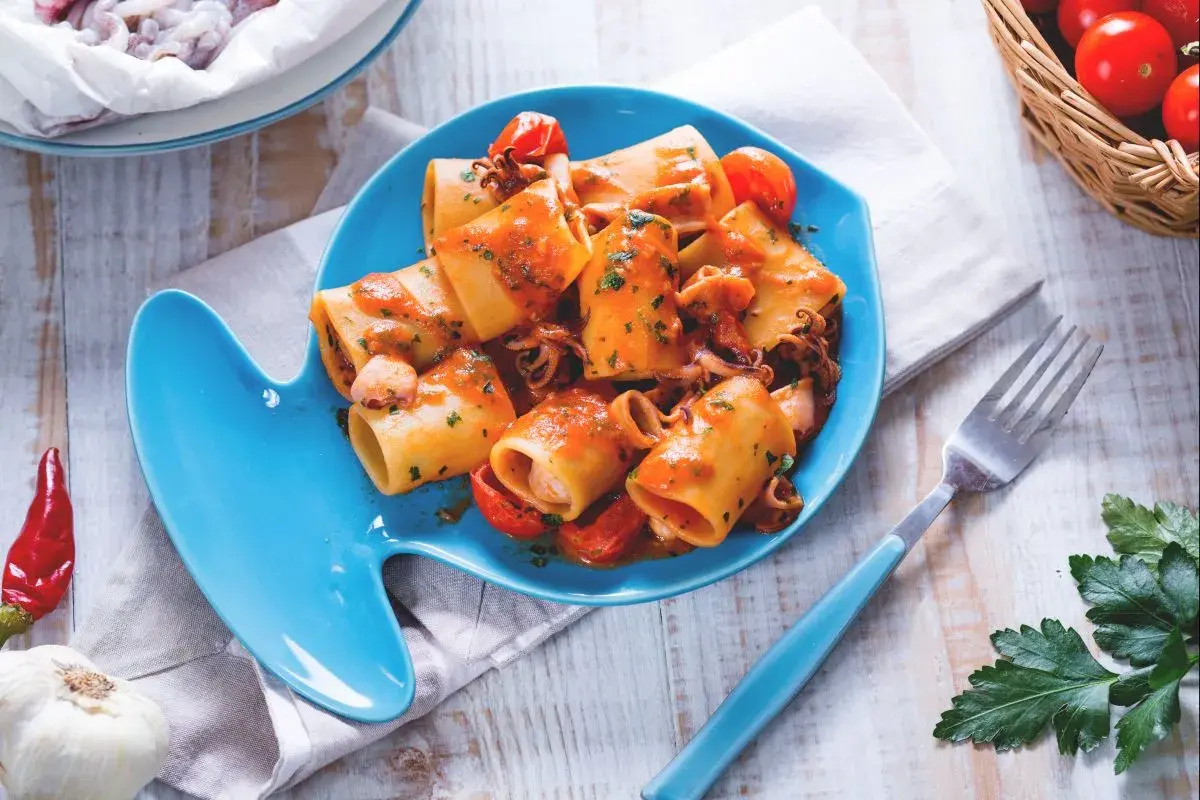 Piatto di pasta con calamaretti, pomodorini e prezzemolo su un piatto a forma di pesce blu.