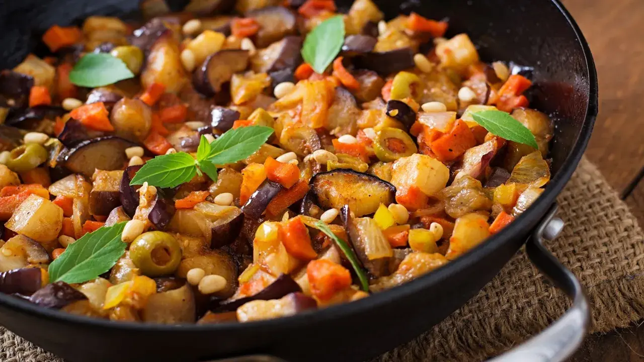 Caponata di verdure in padella, con melanzane, carote, olive e pinoli, guarnita con foglie di basilico fresco.