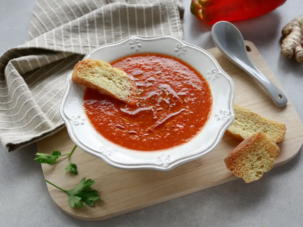 Una ciotola di crema di peperoni, guarnita con crostini e un filo d'olio. Accanto, un cucchiaio, un peperone rosso e zenzero.