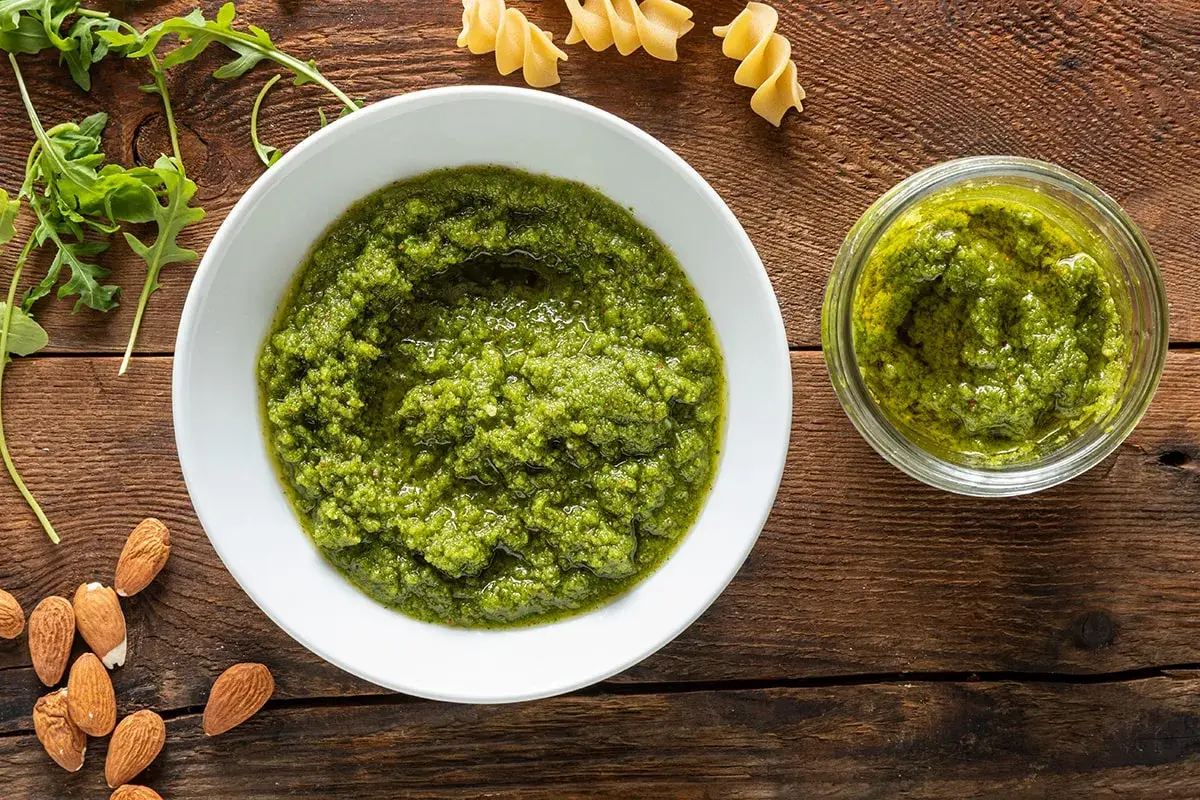 Pesto di rucola e mandorle in una ciotola bianca, accanto a un vasetto pieno, con mandorle e rucola fresca sparse sul tavolo di legno.