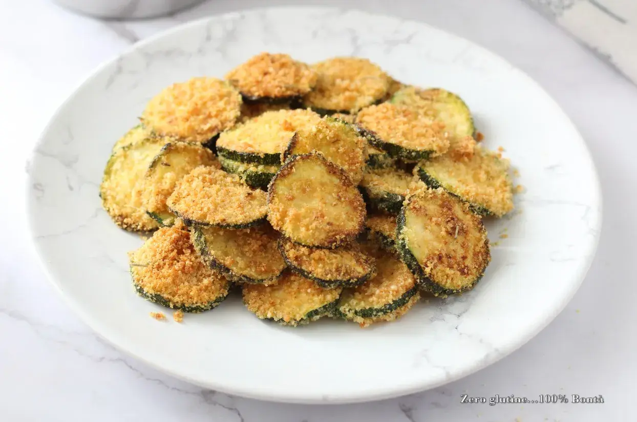 Fettine di zucchine impanate e cotte in friggitrice ad aria, dorate e croccanti, servite su un piatto bianco con venature grigie.