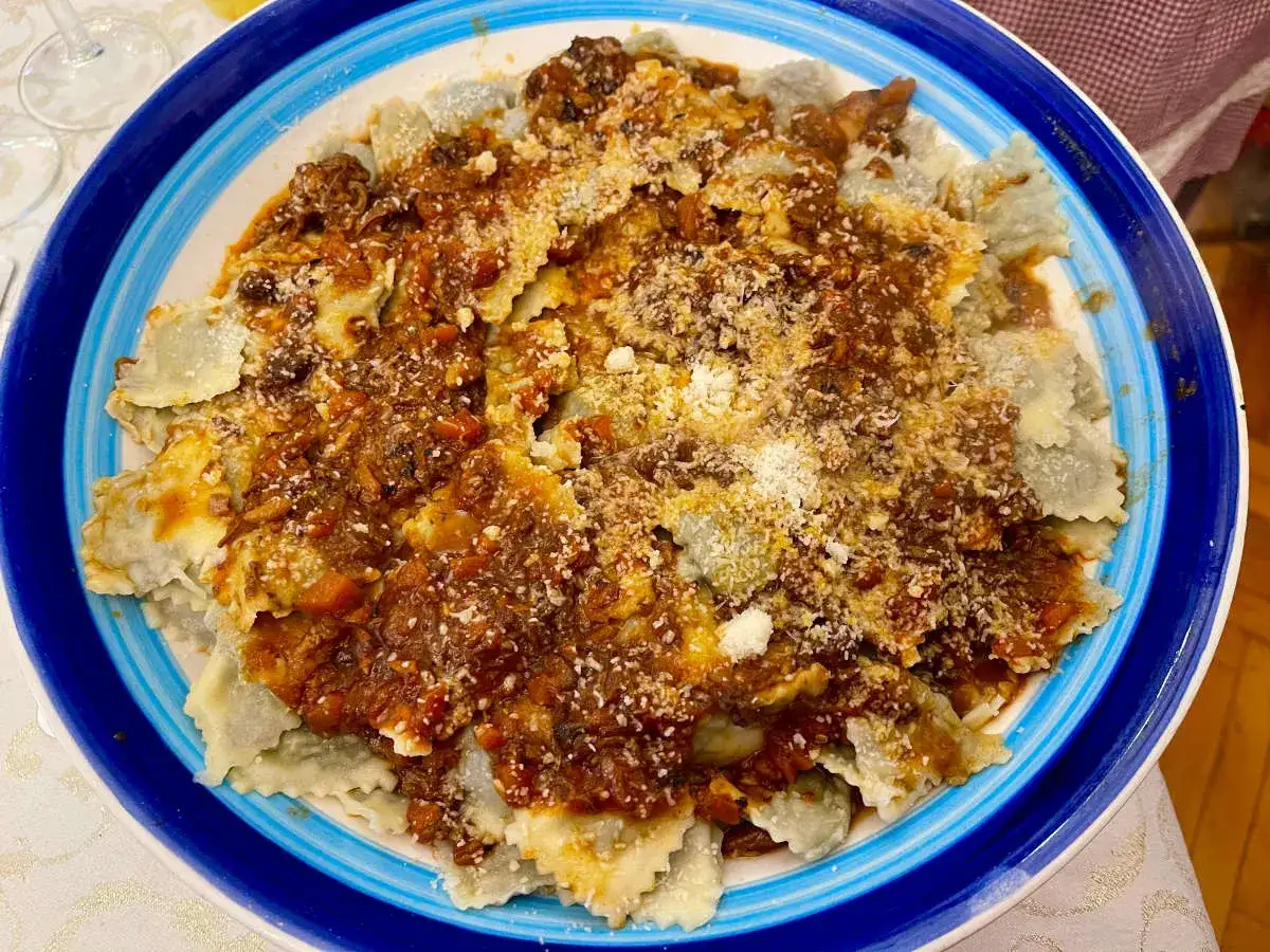 Piatto di ravioli alla genovese con sugo ricco e abbondante parmigiano grattugiato, serviti in un piatto blu e bianco.