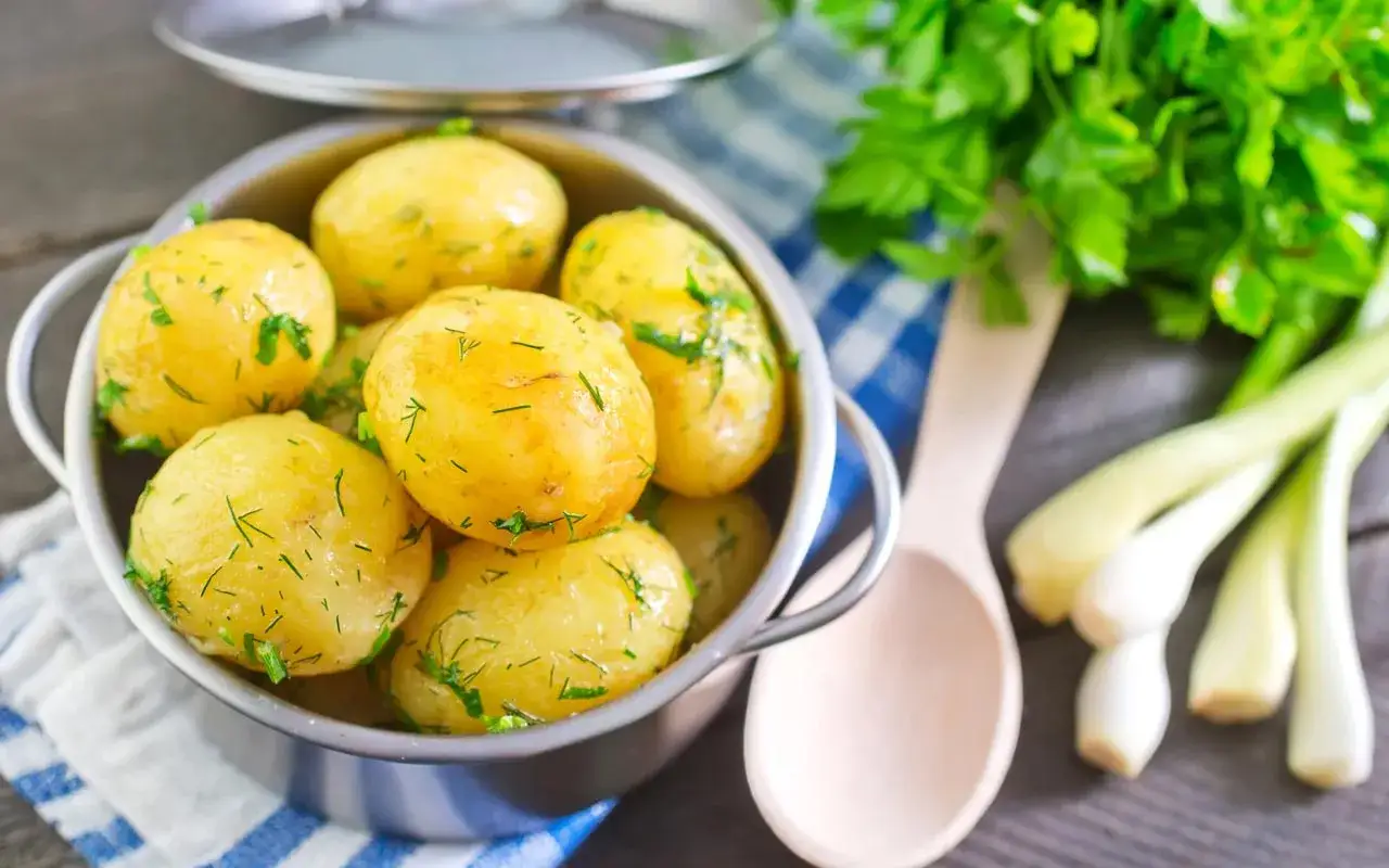 Patate novelle bollite, condite con prezzemolo fresco e un filo d'olio. Accanto, cipollotti e un cucchiaio di legno.