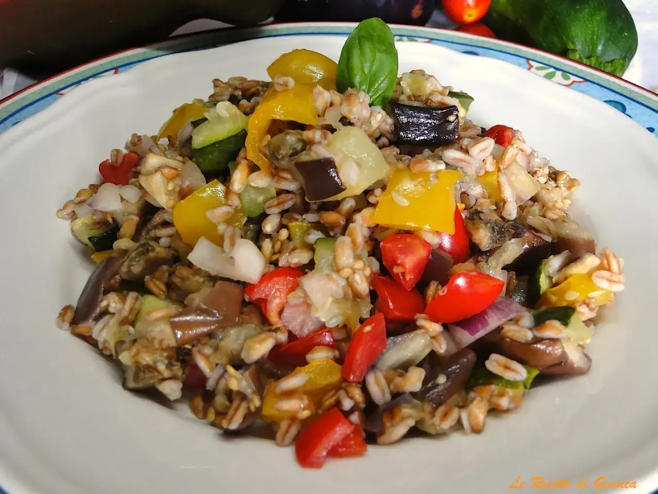 Un colorato farro con verdure miste, tra cui melanzane, zucchine, peperoni gialli e pomodorini, guarnito con una fogliolina di basilico.