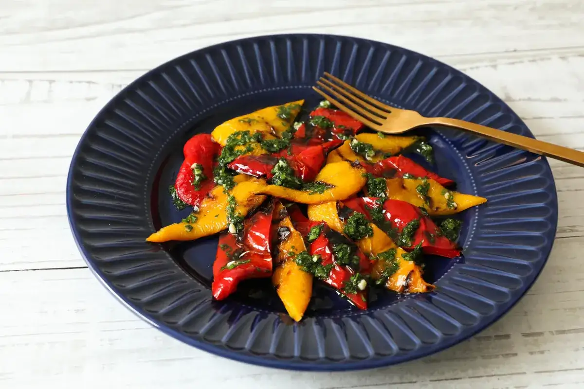 Peperoni in padella, rossi e gialli, conditi con erbe aromatiche, su un piatto blu scuro.