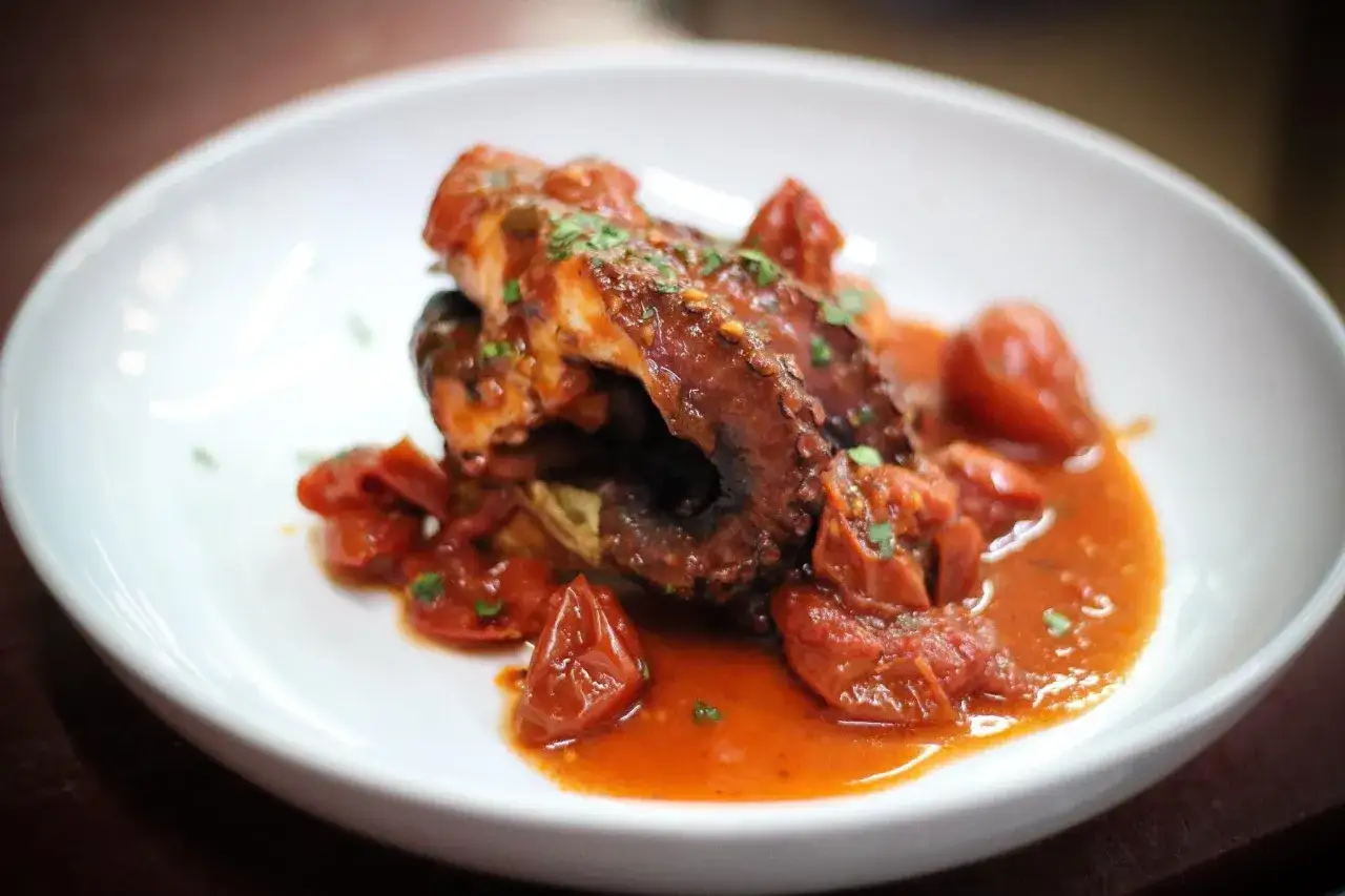 Un saporito polpo alla luciana, tenero e succulento, servito in un piatto bianco con pomodorini e un ricco sugo rosso.
